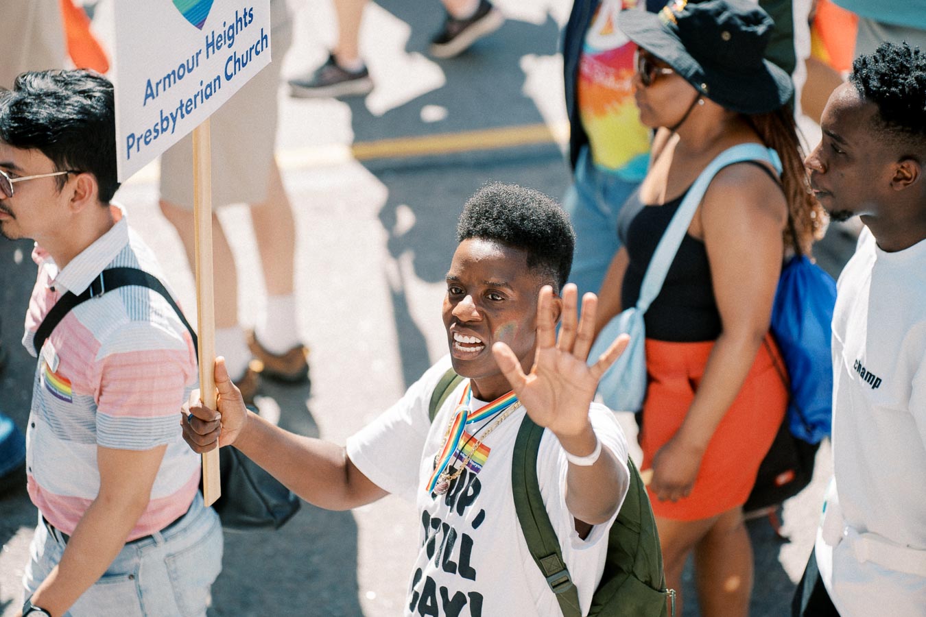 Participants in a parade, one holding a sign for Armour Heights Presbyterian Church, celebrating with rainbow accessories.