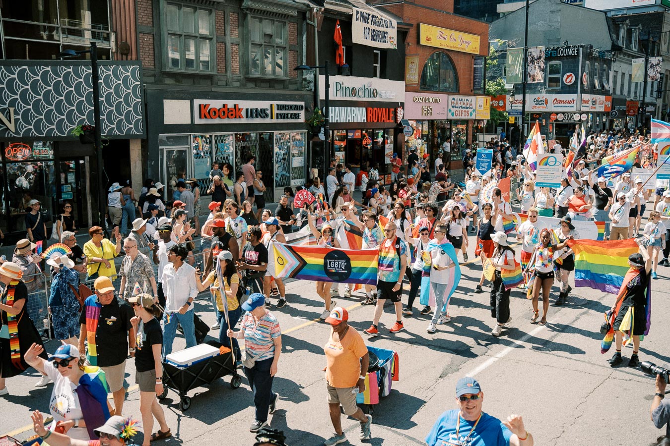 Participants in a lively LGBTQ+ pride parade march down a city street, proudly displaying rainbow flags and banners. The street is lined with various shops and spectators.