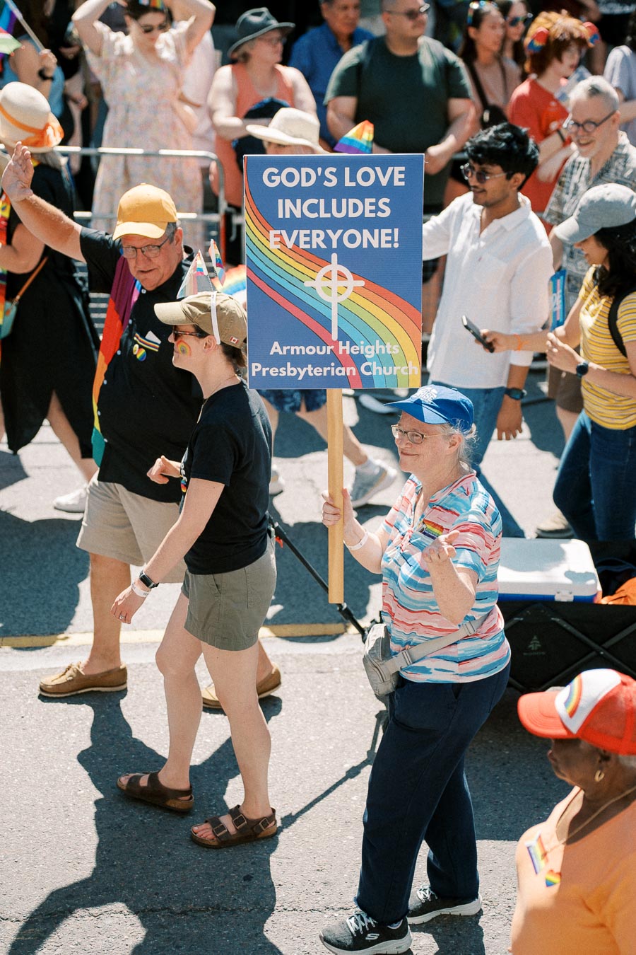 Participants in a parade hold a sign promoting inclusivity and love from Armour Heights Presbyterian Church, featuring a rainbow design.