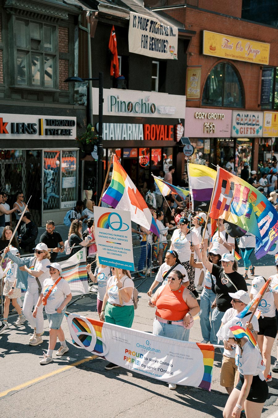 Participants in a pride parade march with rainbow flags and banners promoting fairness in Ontario public services, set against a backdrop of city shops and onlookers.