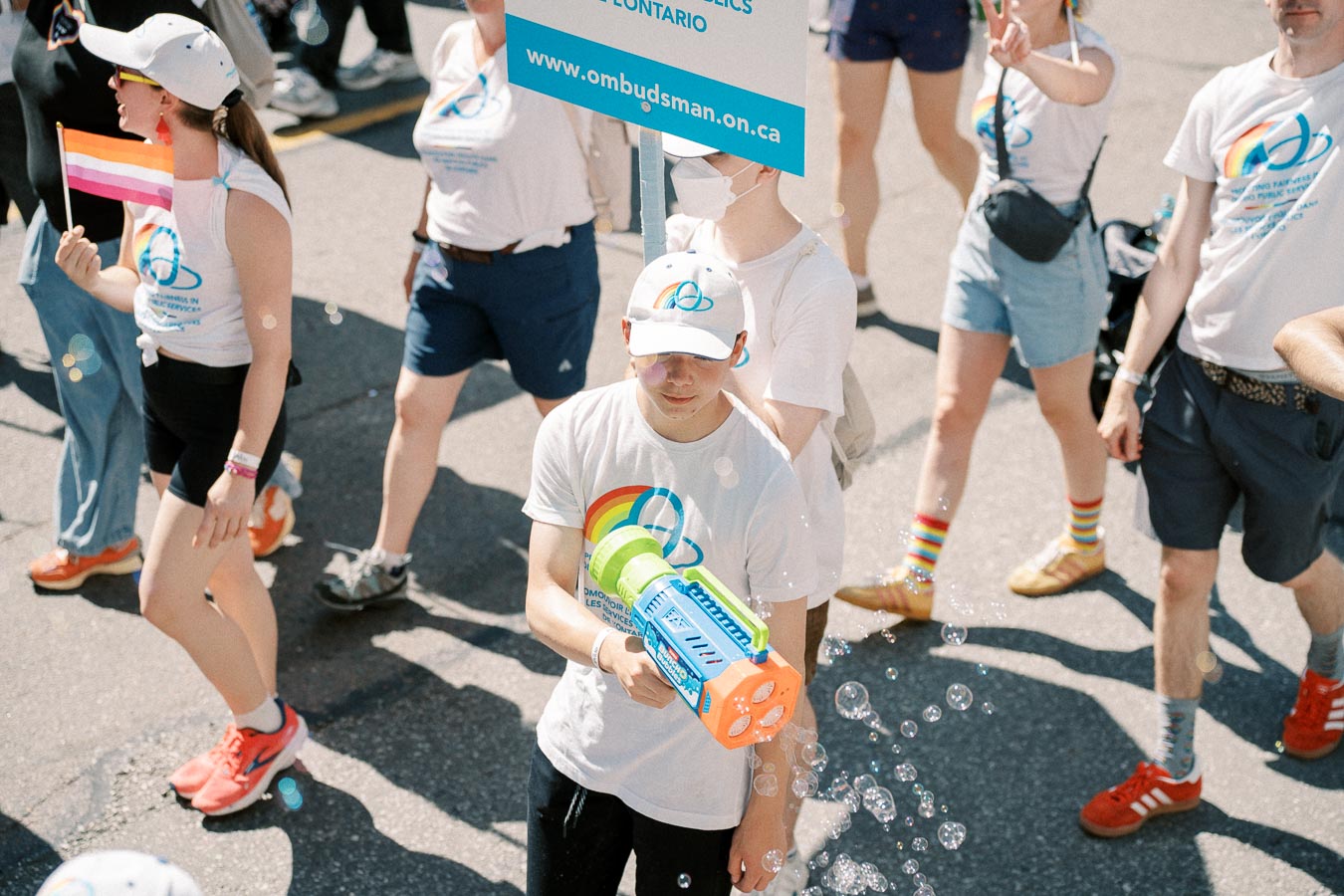 Participants in a parade wearing white shirts with rainbow designs, holding a sign and a bubble gun, celebrating diversity and inclusion.