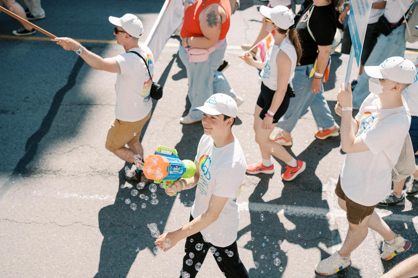 Participants in a parade wearing matching white t-shirts and caps, with one person joyfully using a bubble gun.