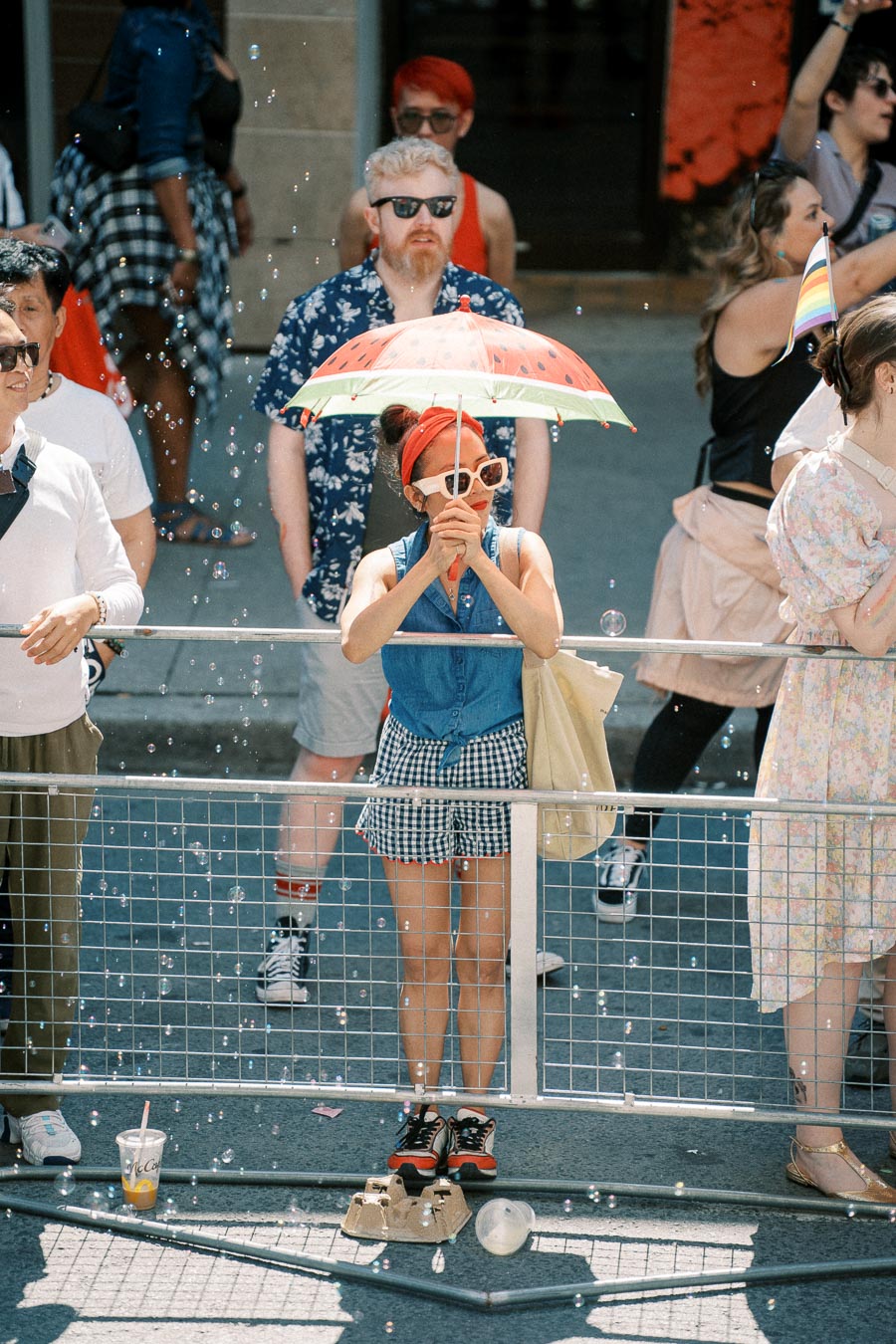 A person stands behind a barrier at an outdoor event, holding a watermelon-themed umbrella. They are surrounded by bubbles and dressed in a denim top and checkered shorts, with a red headband and sunglasses.