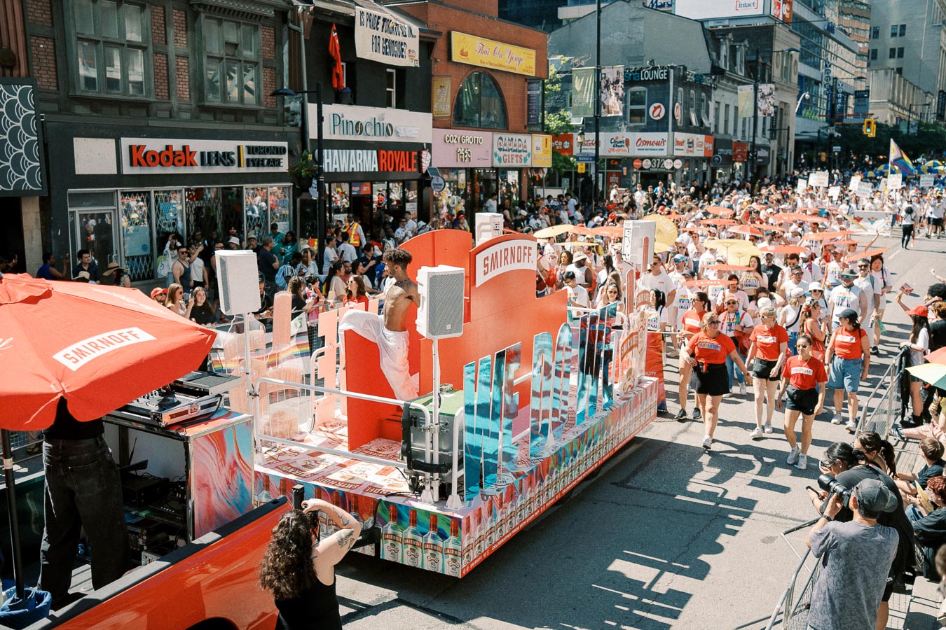 A colorful parade featuring a Smirnoff float and participants marching down a bustling city street, with spectators lining the sidewalks.