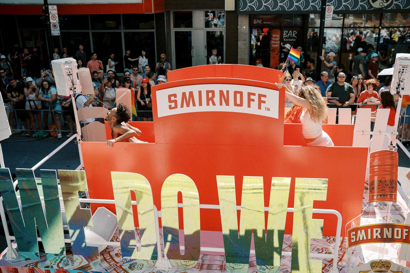 A vibrant parade float featuring a large Smirnoff sign, with people celebrating and waving a rainbow flag, surrounded by a cheering crowd.