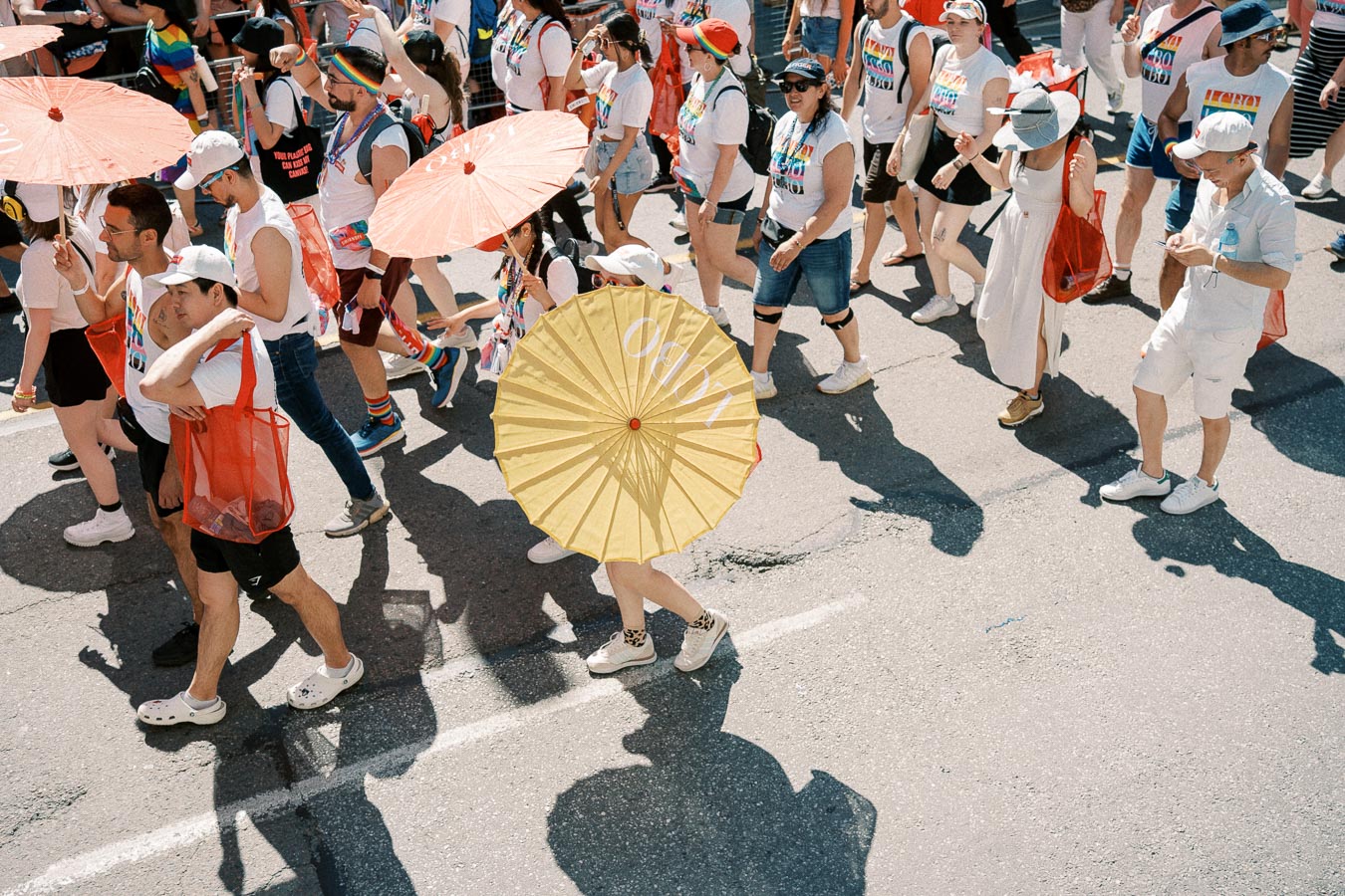 Participants in a pride parade march down the street, holding colorful parasols and wearing rainbow-themed attire.