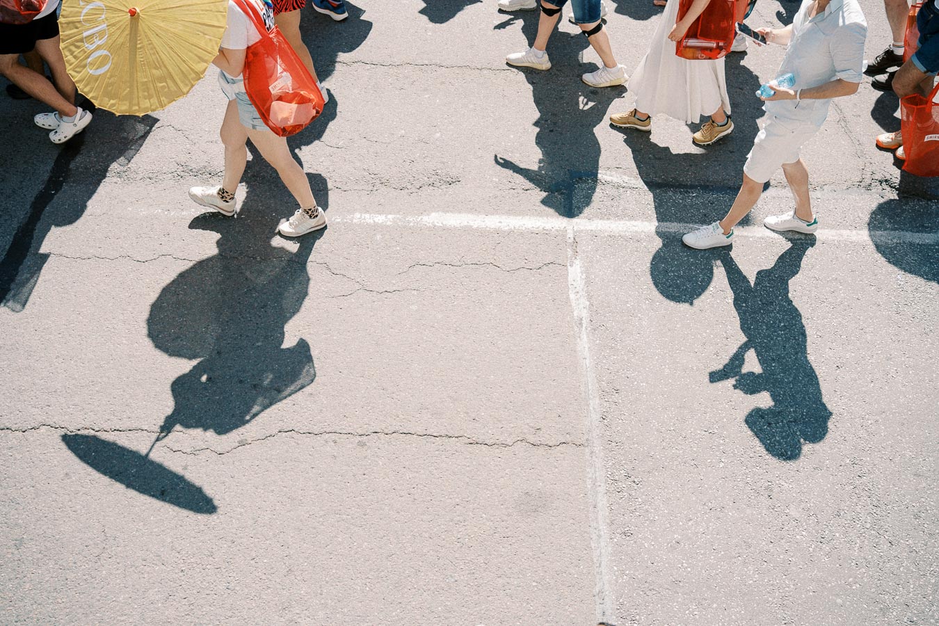 Aerial view of pedestrians walking on a street, casting distinct shadows. A person is holding a yellow parasol, adding a splash of color to the scene.