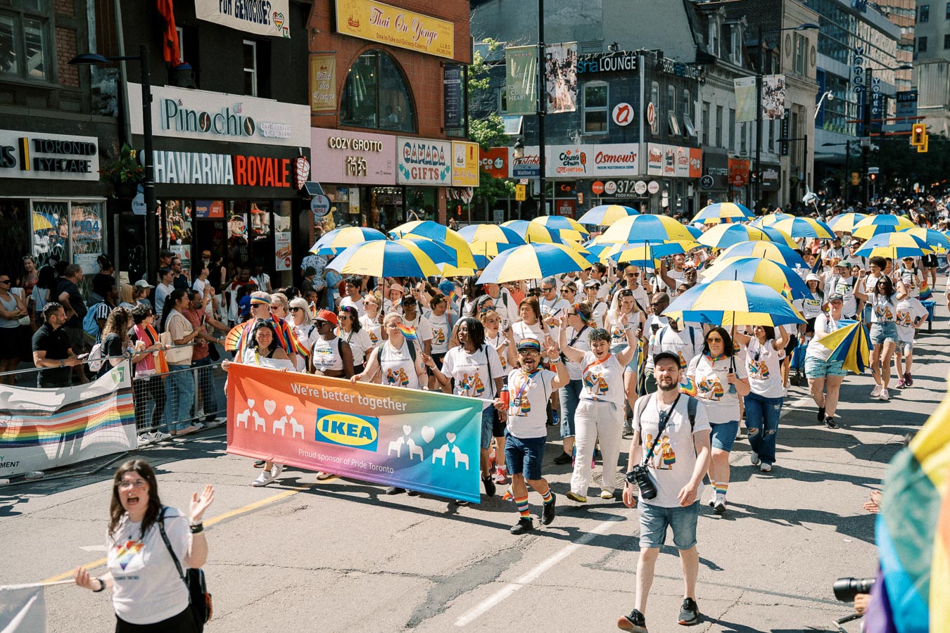 Participants in a parade march with blue and yellow umbrellas, holding a banner that reads 'We're better together' with the IKEA logo, as spectators watch from the side.