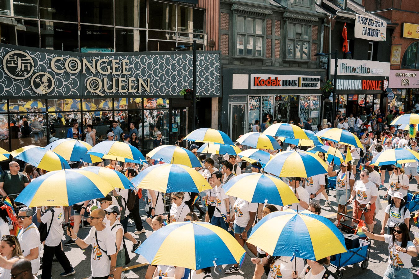 Parade participants walk with blue and yellow umbrellas past Congee Queen restaurant.