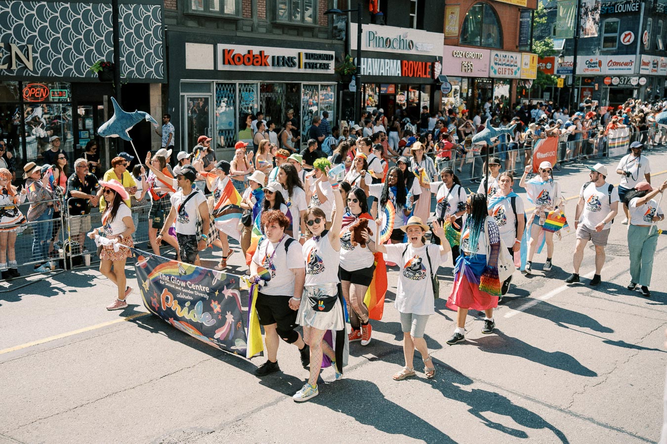 Participants in a pride parade march down a city street, carrying a banner and wearing matching shirts, surrounded by cheering spectators.