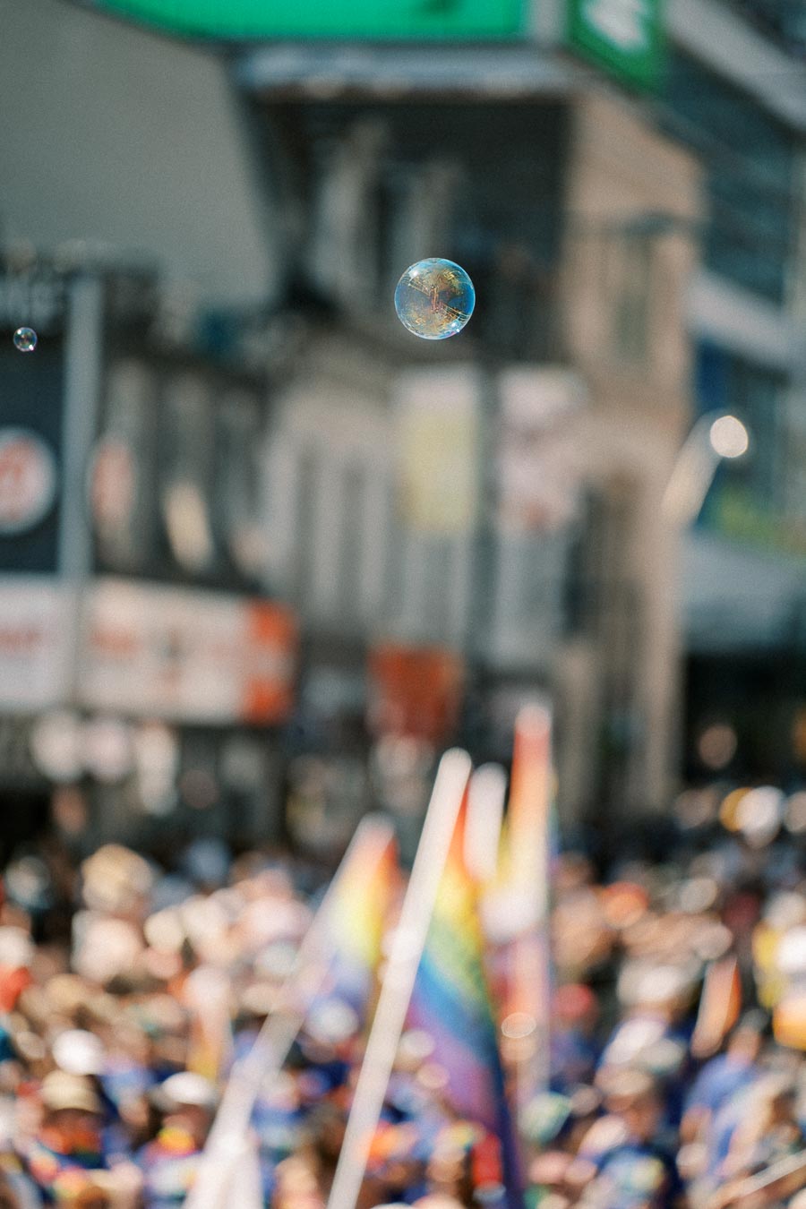 A soap bubble floats above a vibrant crowd at a parade, with rainbow flags visible in the background, creating a festive atmosphere.