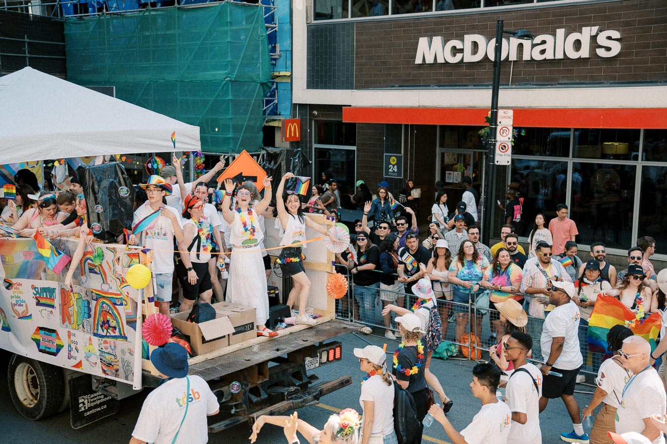 A lively parade with participants on a decorated float and spectators on the street, celebrating with rainbow flags and colorful attire in front of a McDonald's.