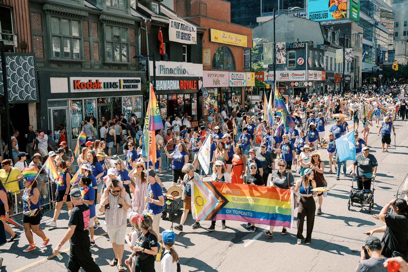 Participants in a Toronto parade celebrate with rainbow flags and a banner for 'Proud Elementary Teachers of Toronto'.