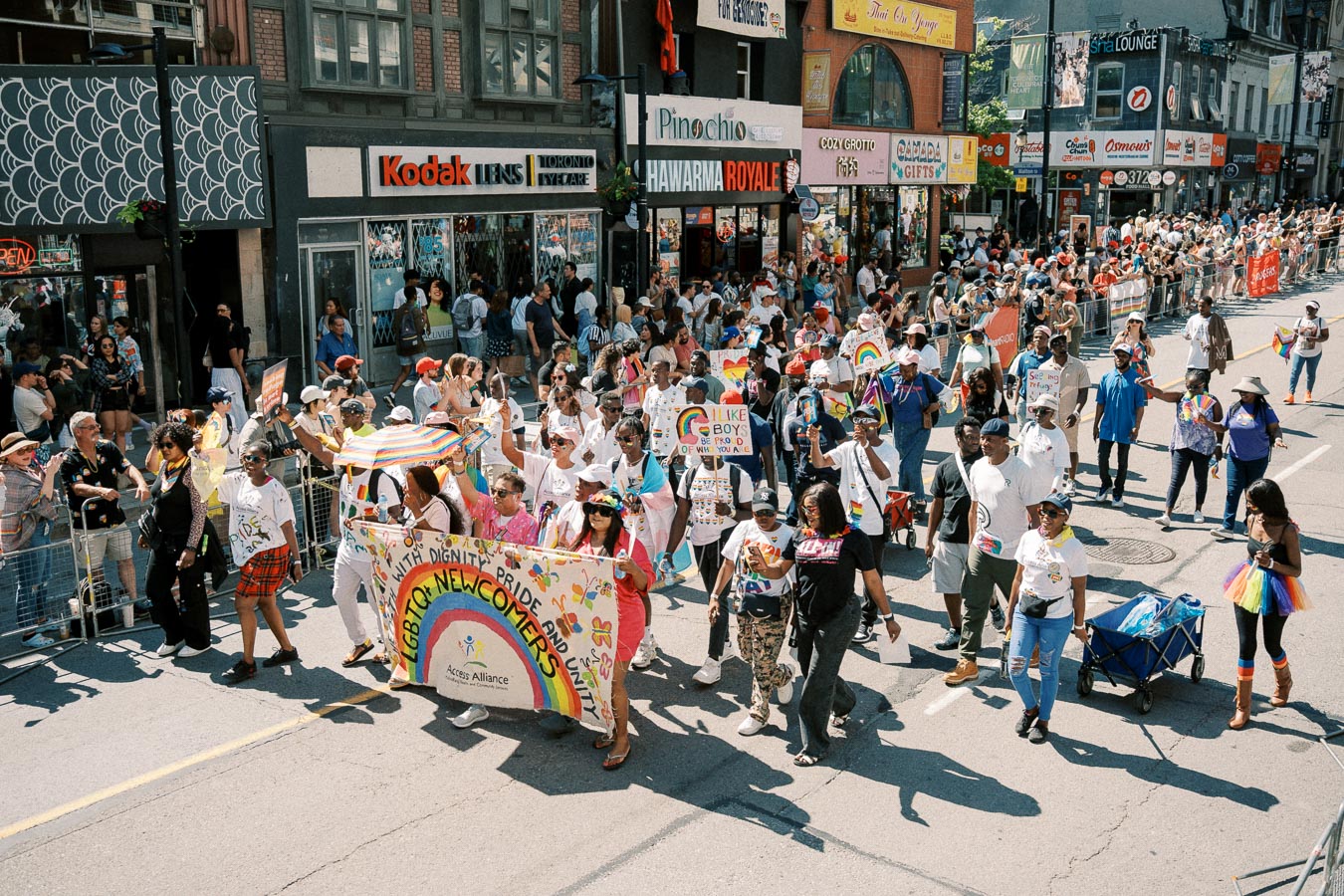 Participants in a lively LGBTQ+ pride parade march down a city street, holding a colorful banner and rainbow flags, while spectators watch from the sidelines.