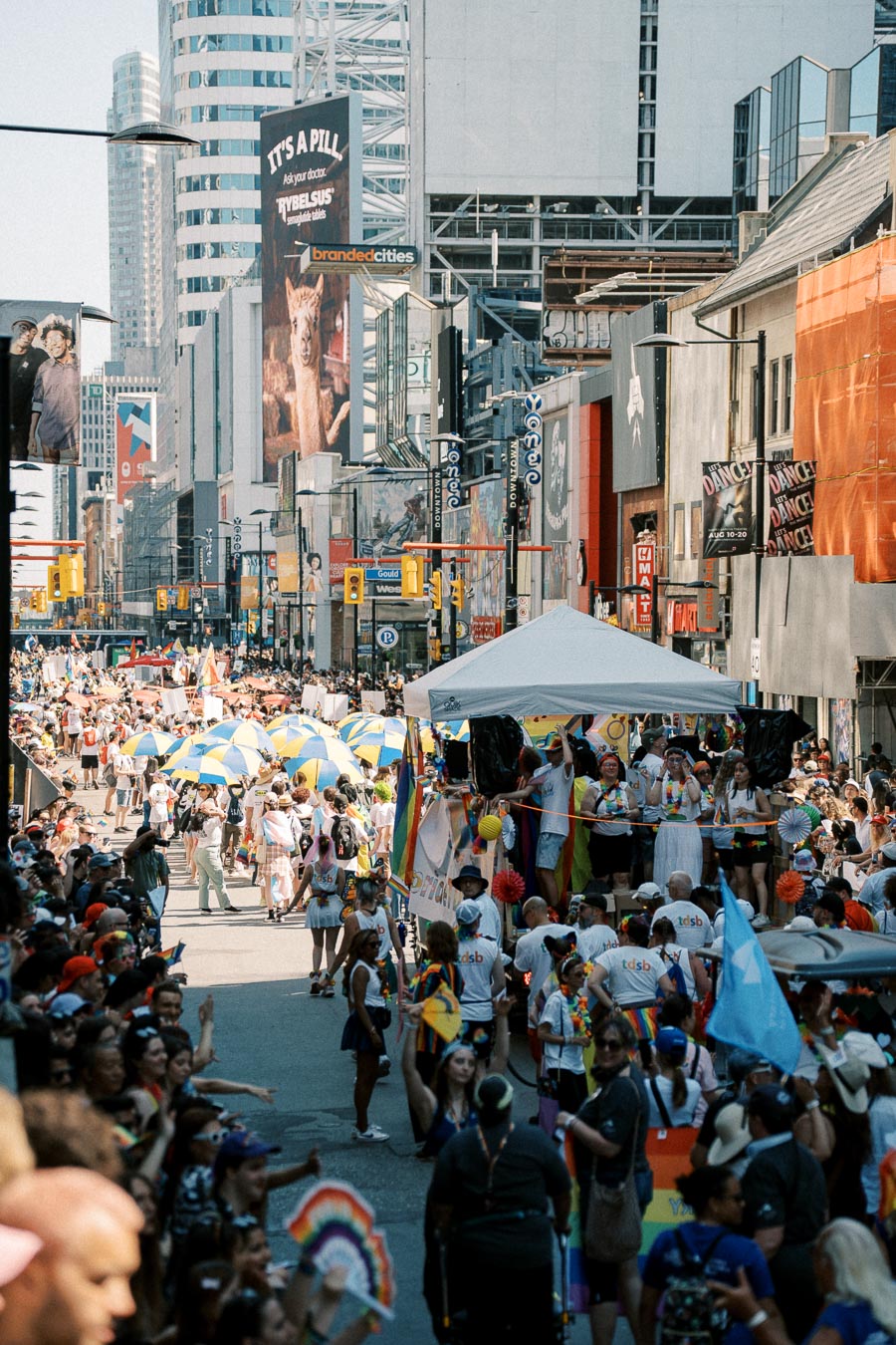 A lively street parade featuring colorful umbrellas and rainbow flags, set against a backdrop of urban buildings and billboards.