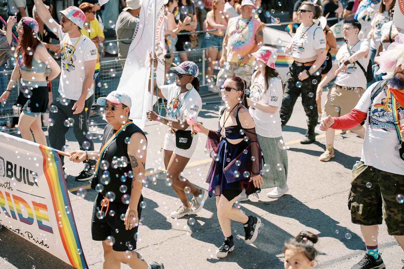 Participants in a pride parade march joyfully, surrounded by bubbles and wearing colorful attire and accessories.