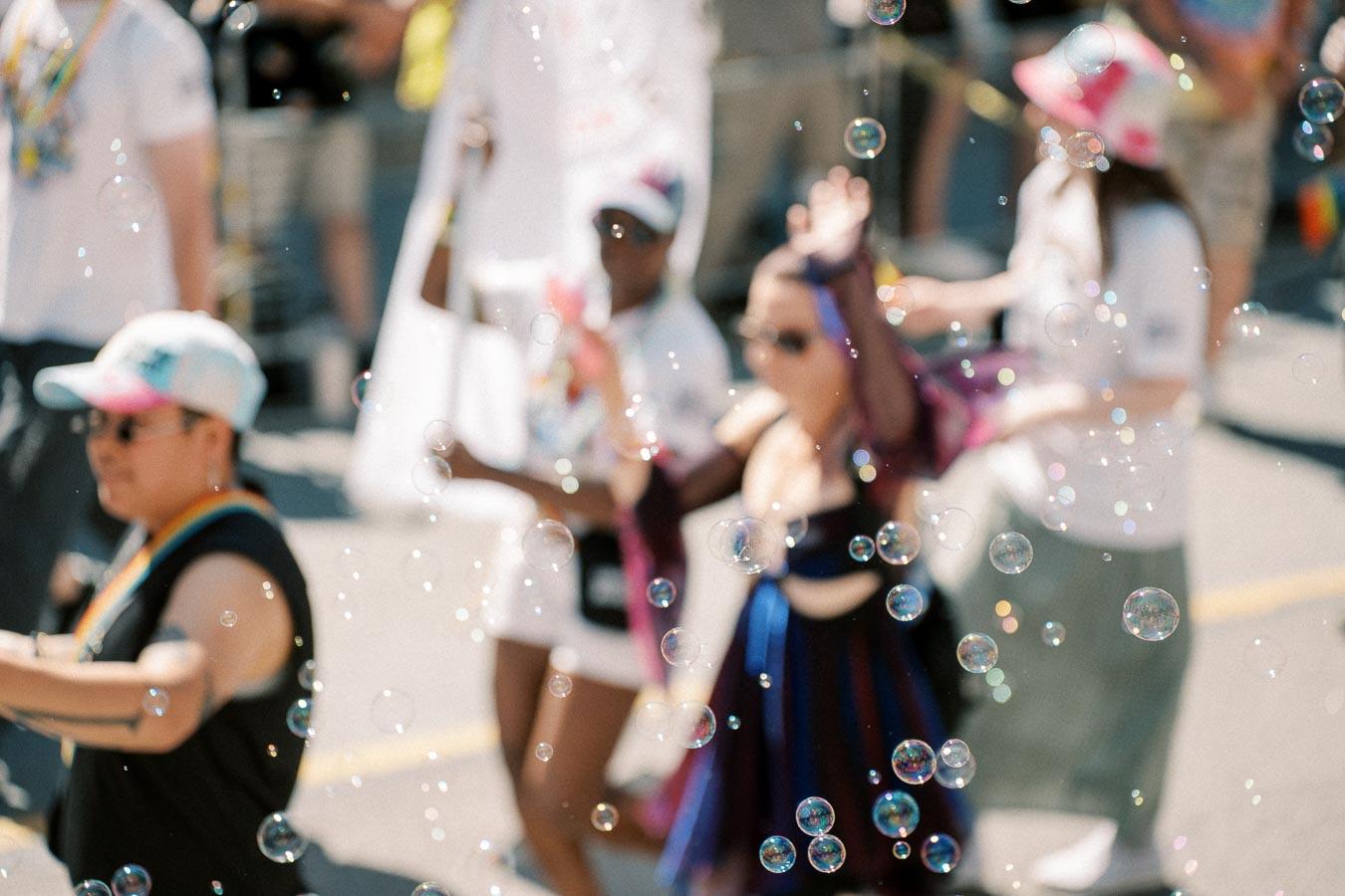 A vibrant parade scene with people in colorful attire and bubbles floating in the foreground, creating a festive atmosphere.
