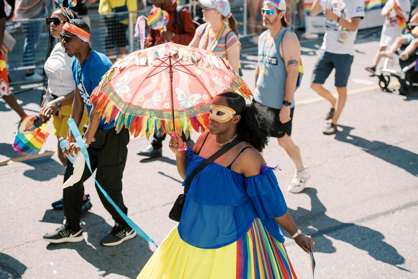 Participants in a vibrant parade, featuring colorful outfits and accessories, including a person holding a decorative umbrella and wearing a mask.