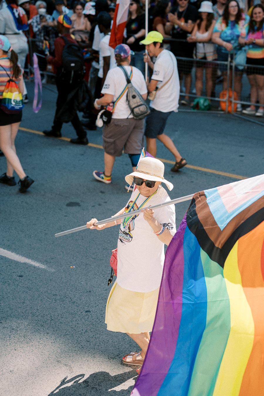 A participant at a pride parade holds a rainbow flag, wearing a sun hat and sunglasses, surrounded by a crowd.