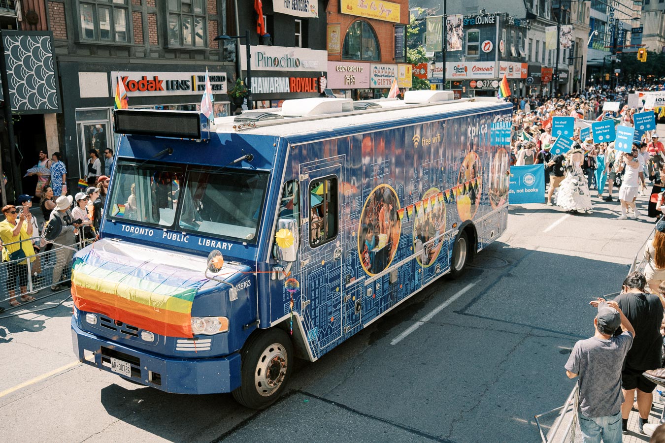 A Toronto Public Library bookmobile adorned with rainbow flags participates in a lively parade, surrounded by a crowd of spectators and marchers holding signs.