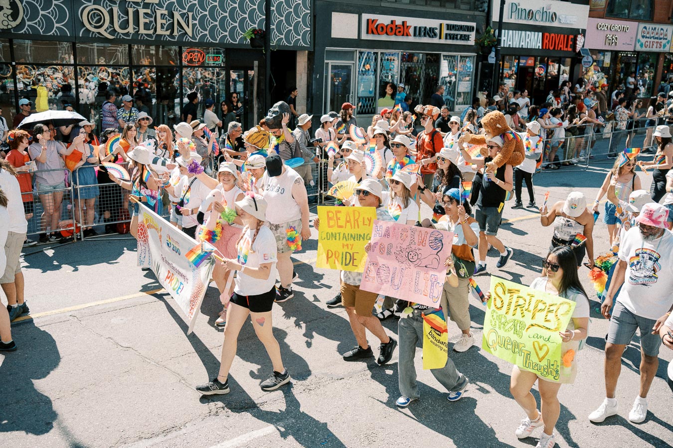 Participants in a pride parade march down a city street, holding vibrant signs and wearing rainbow-themed accessories. Spectators line the sidewalks, cheering and taking photos.