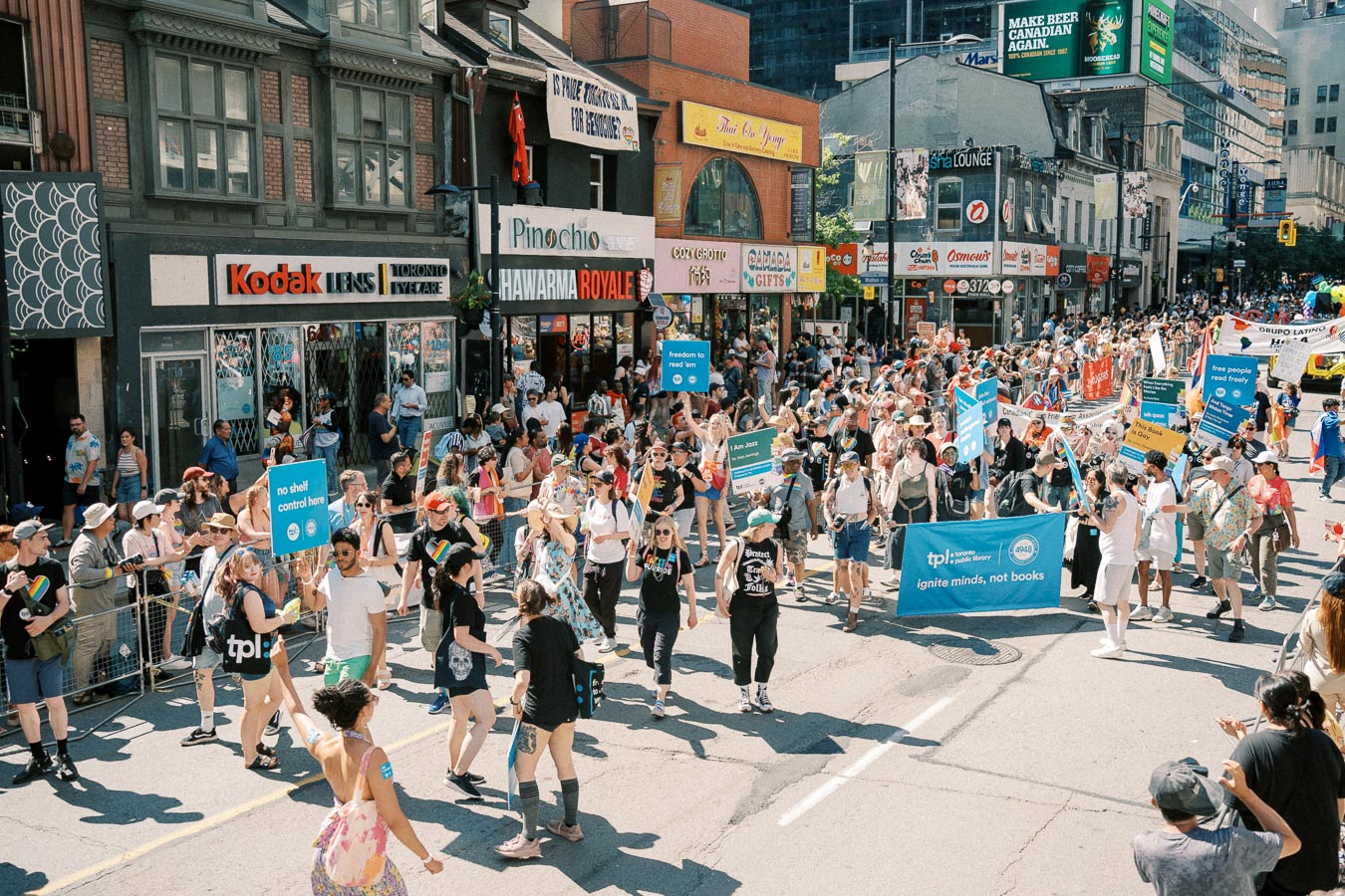 A vibrant parade featuring diverse participants holding signs and banners, celebrating on a city street lined with shops.