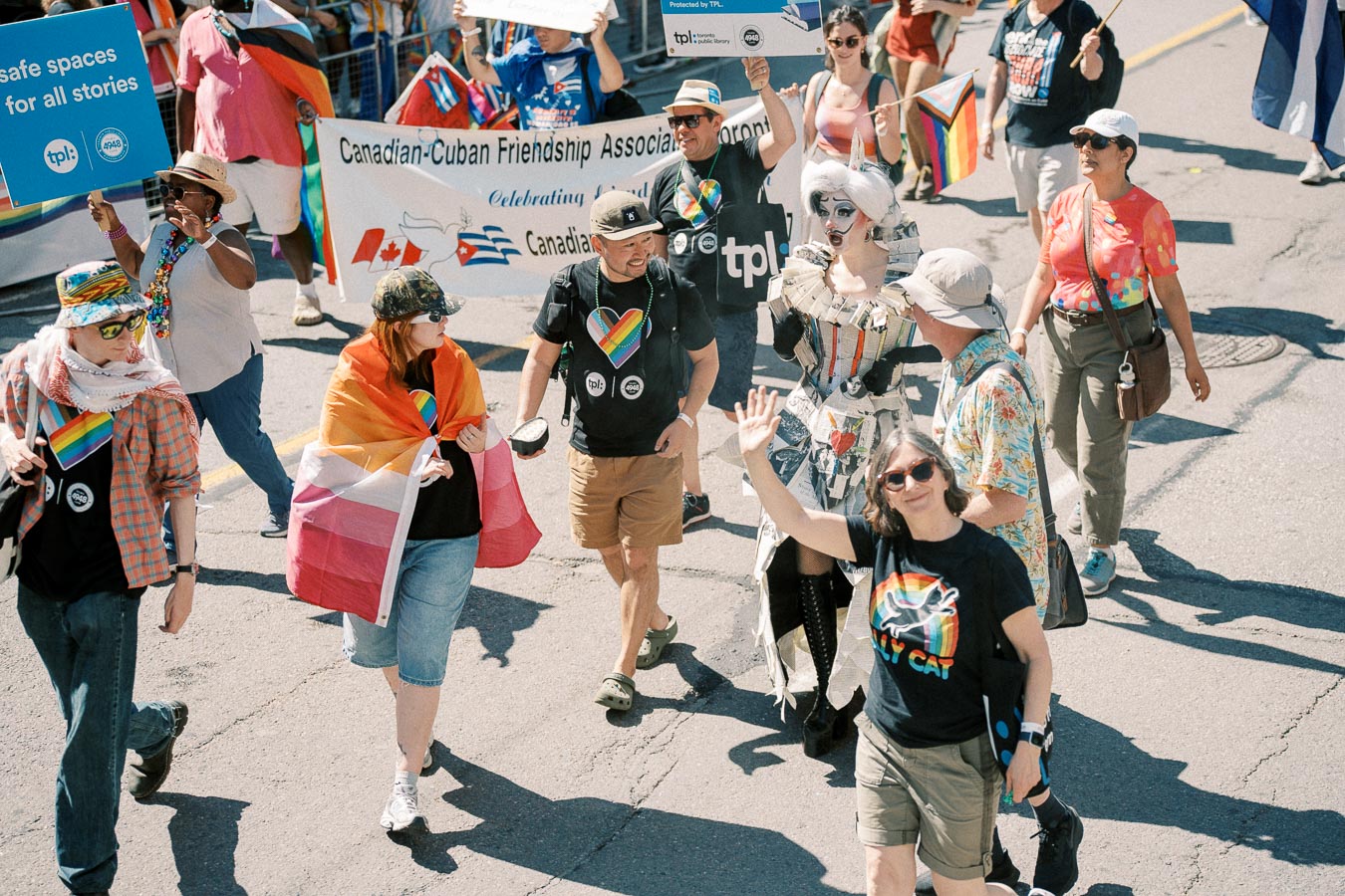 Participants in a parade celebrate diversity and inclusion, showcasing rainbow flags and signs promoting safe spaces. The Canadian-Cuban Friendship Association Toronto banner is visible.