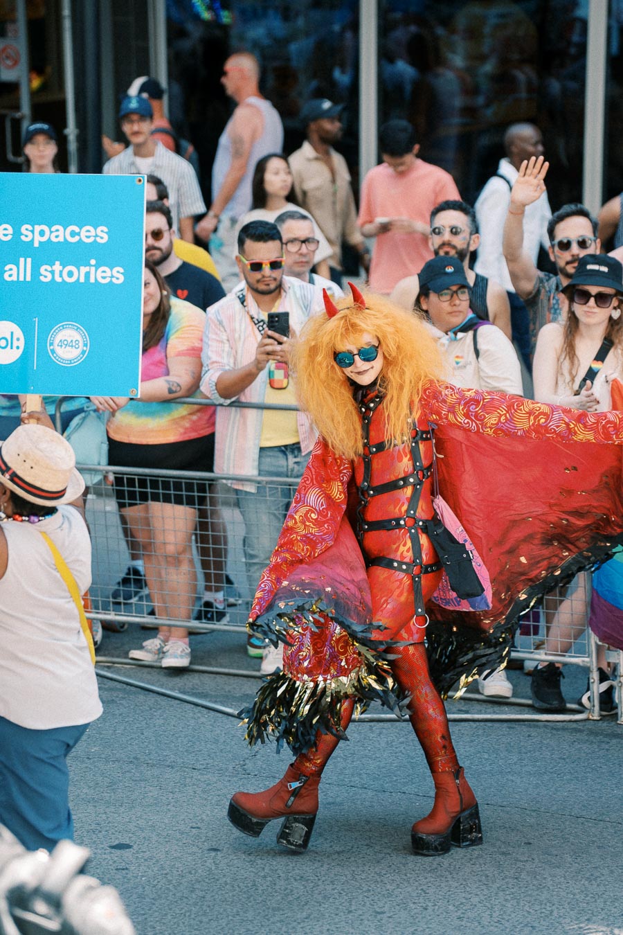 A colorful parade participant in a vibrant costume with horns and sunglasses, alongside a sign promoting inclusivity.