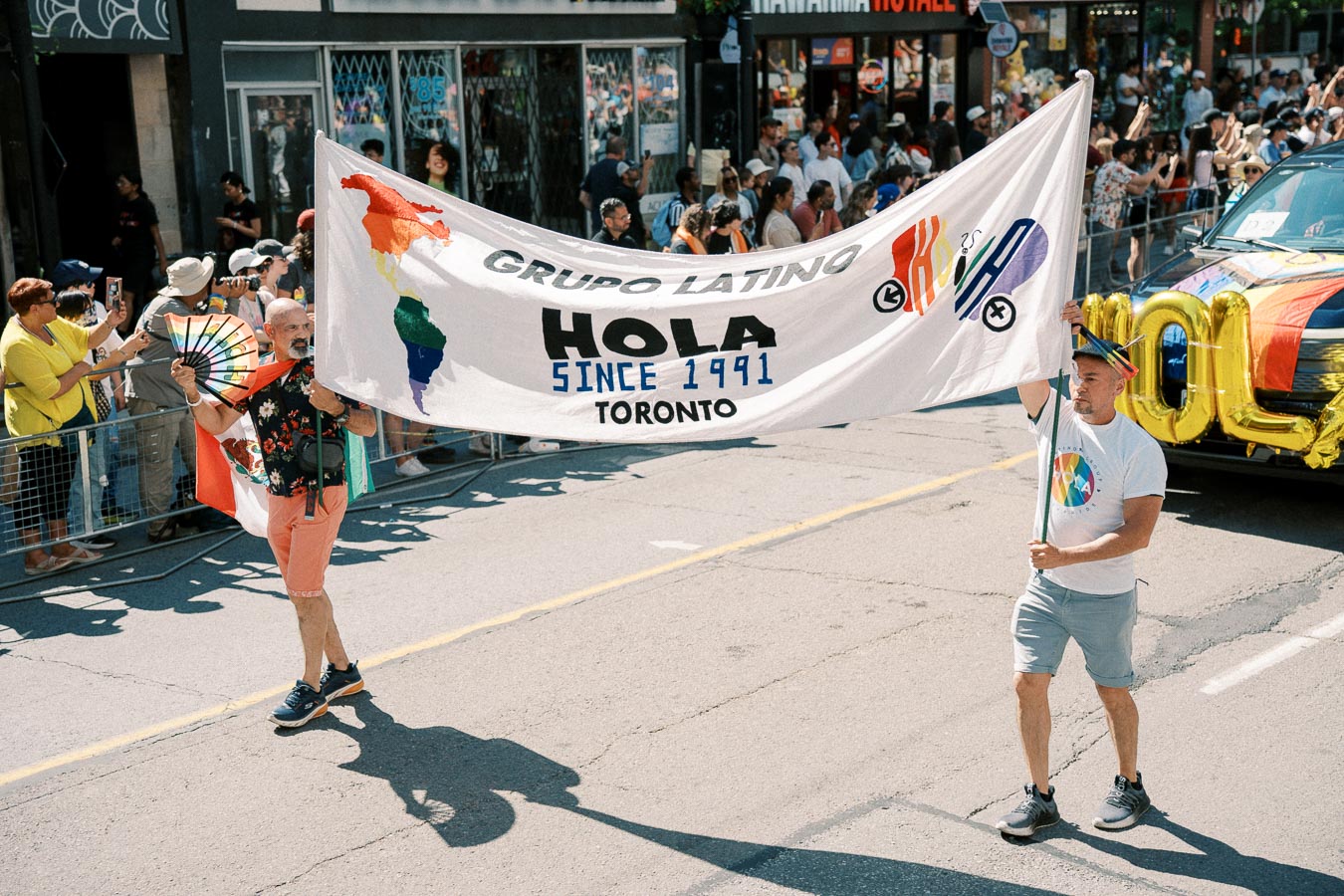 Participants in a Toronto parade hold a banner for Grupo Latino, featuring rainbow colors and the text 'HOLA SINCE 1991'.