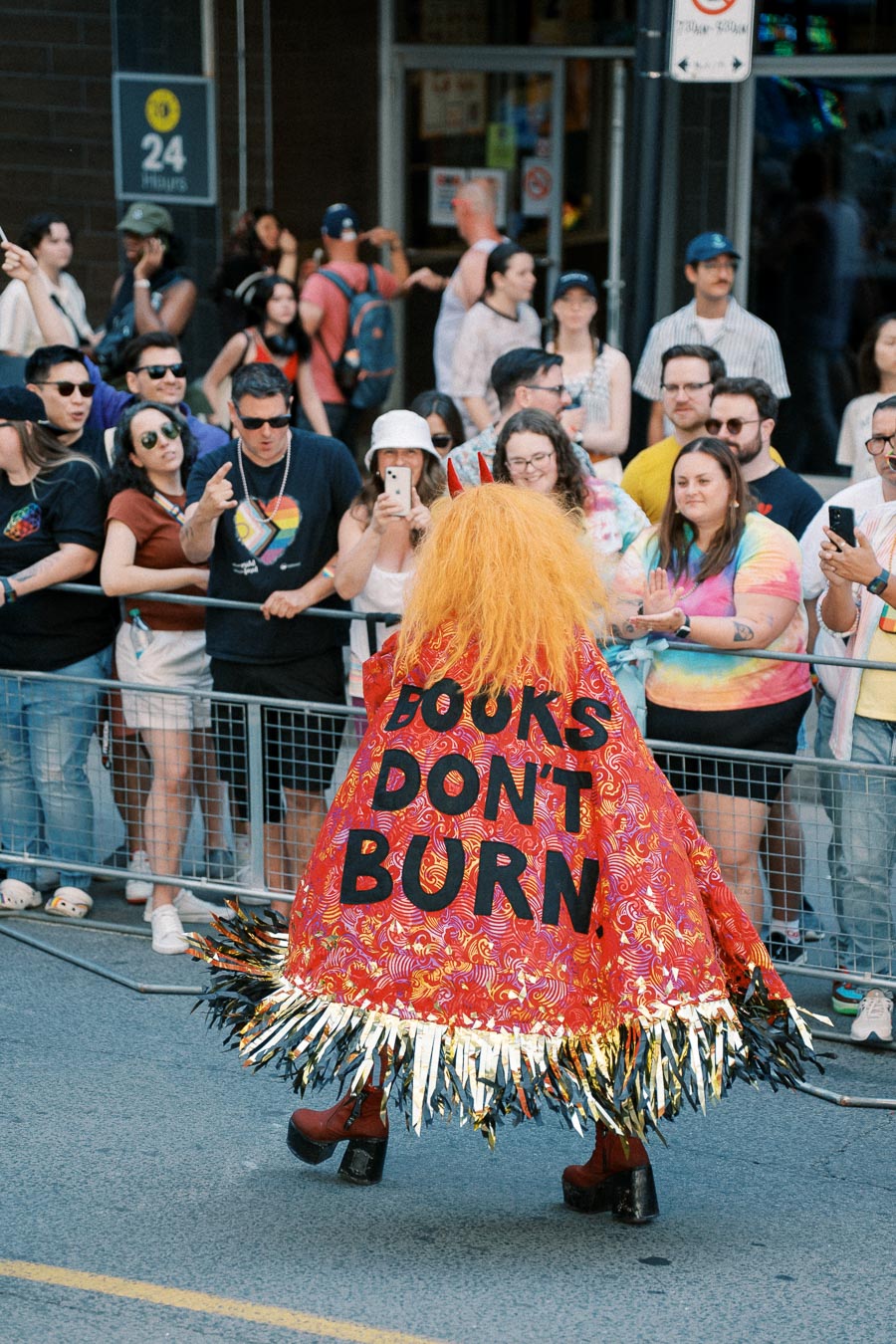 A vibrant parade scene featuring a person in a colorful costume with a cape that reads 'Books Don't Burn', surrounded by an enthusiastic crowd.