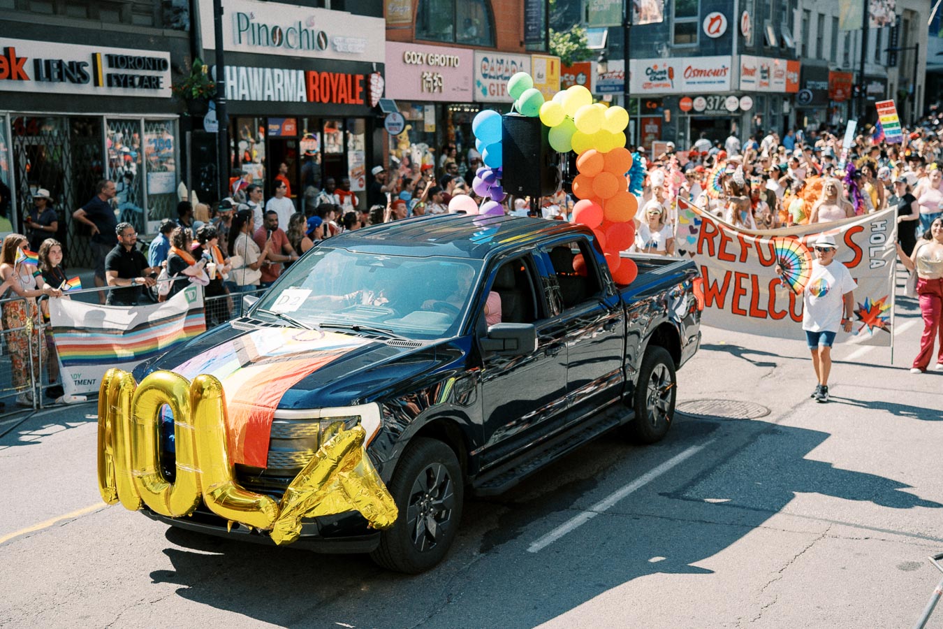 A black pickup truck adorned with rainbow balloons and a flag leads a pride parade, with participants holding a 'Refugees Welcome' banner.