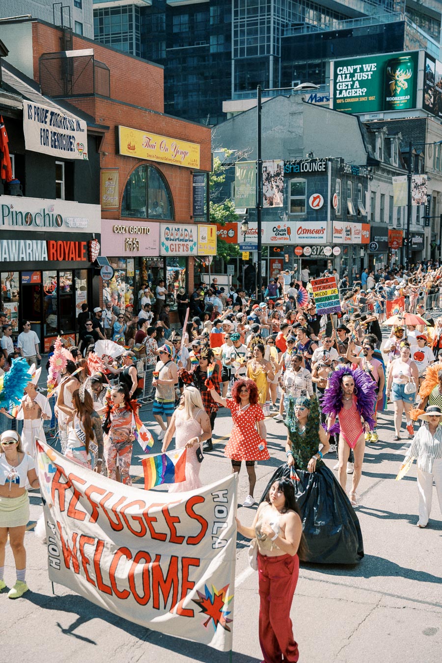 A lively parade featuring participants in colorful costumes and a banner that reads 'Refugees Welcome', set against a backdrop of city shops and spectators.