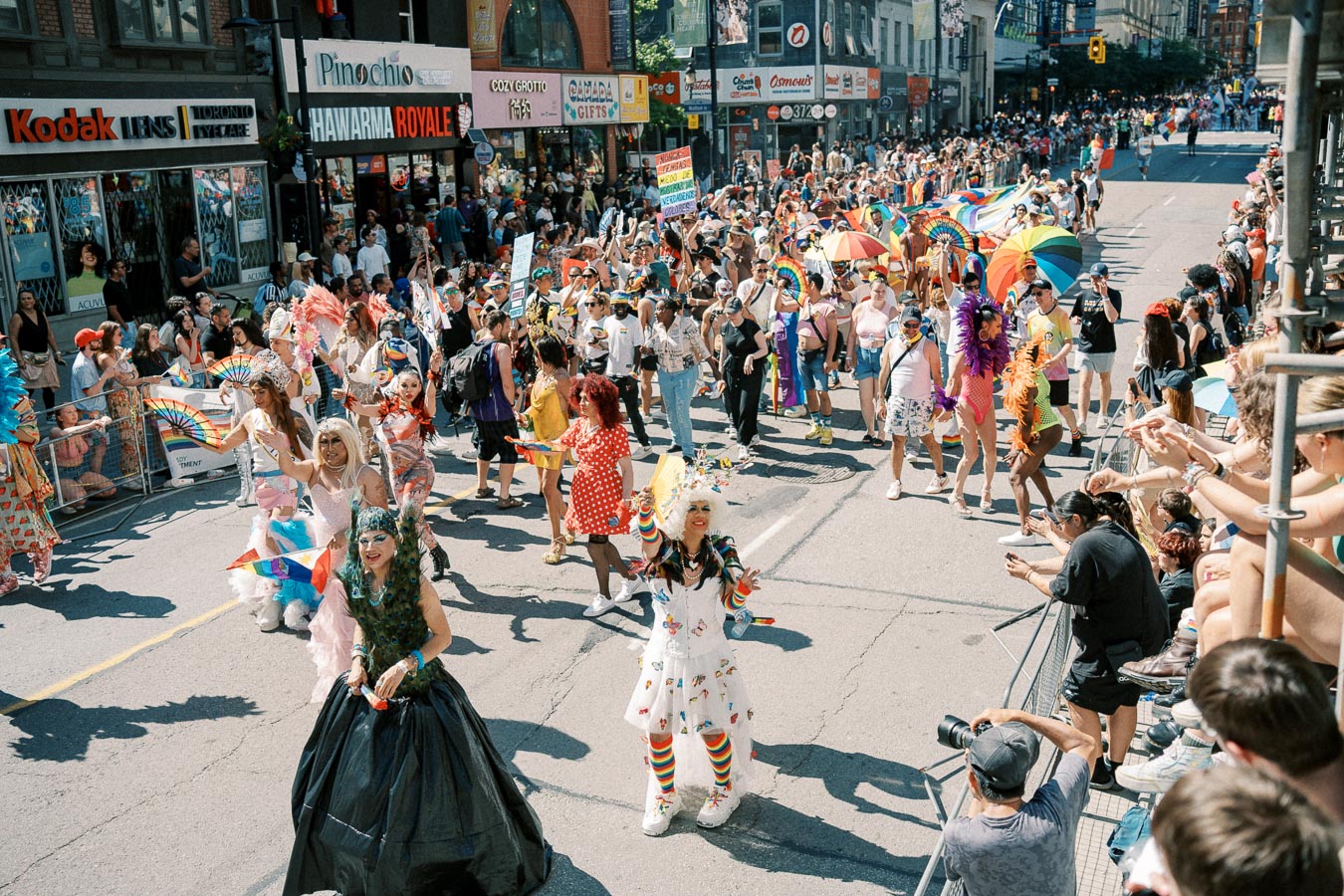 Participants in colorful costumes and rainbow flags march in a lively pride parade, with spectators lining the city street.