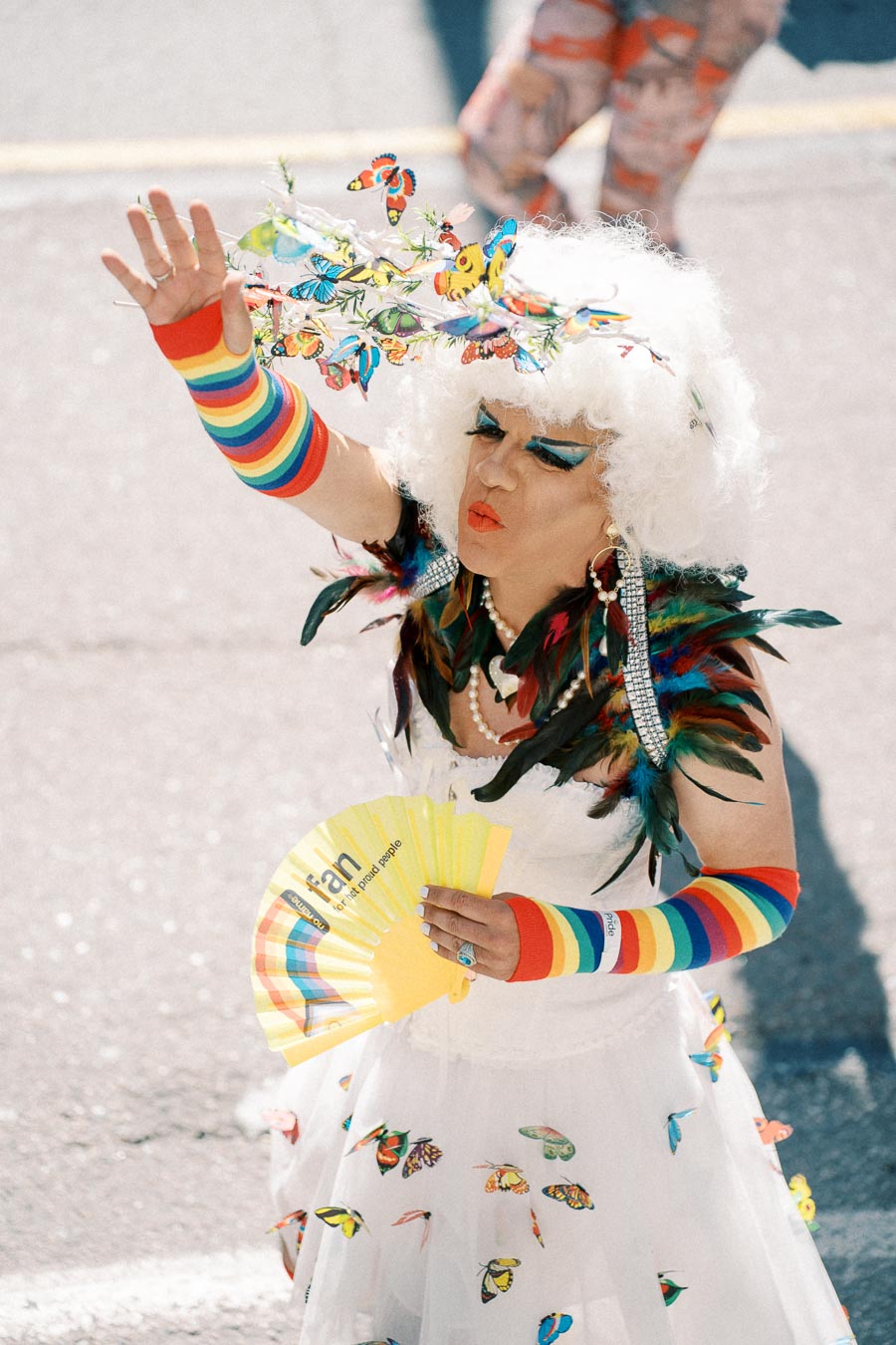 A person dressed in a vibrant costume featuring rainbow arm warmers, a white wig adorned with colorful butterflies, and a white dress with butterfly decorations. They are holding a yellow fan and participating in a parade, showcasing a festive and celebratory atmosphere.
