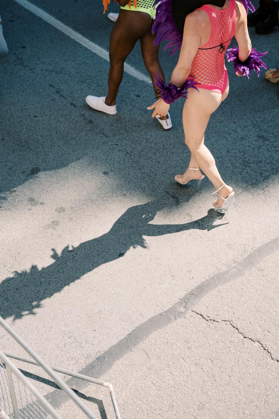 Participants in vibrant costumes walk down a street during a parade, casting distinct shadows on the pavement.