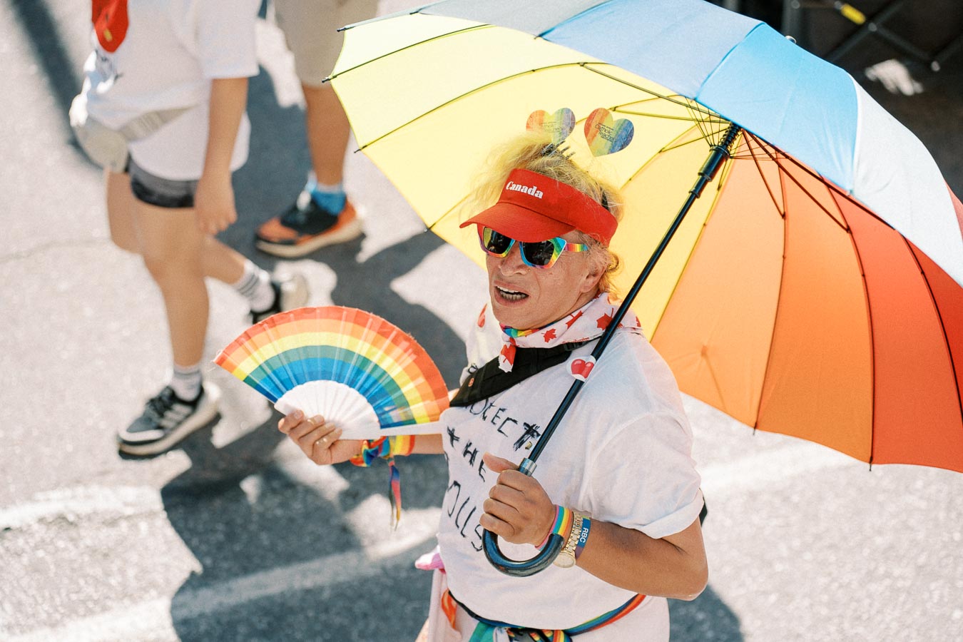 A participant at a pride parade holds a colorful rainbow umbrella and fan, wearing a red visor and rainbow-themed accessories, celebrating diversity and inclusion.