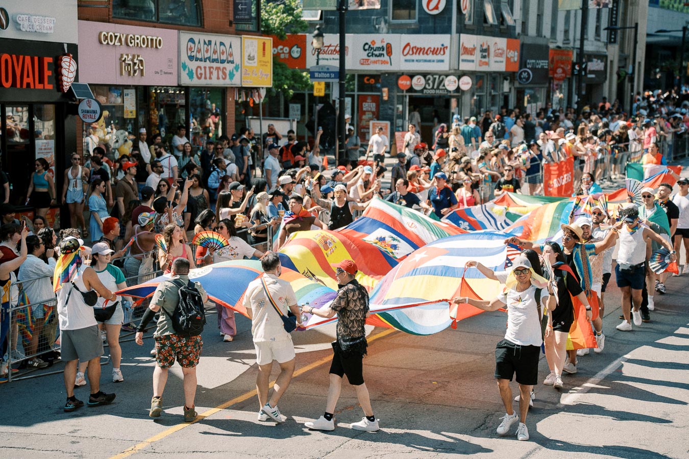 Participants in a lively parade carry a large rainbow flag and various national flags, celebrating diversity and inclusion on a bustling city street.
