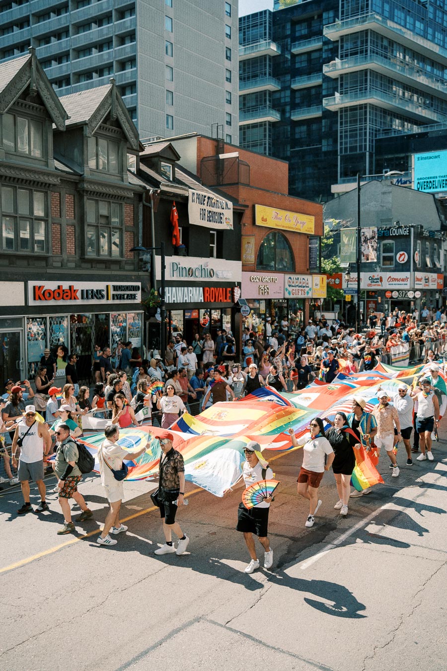 Participants carry a large rainbow flag during a lively street parade, with spectators lining the sidewalks and city buildings in the background.
