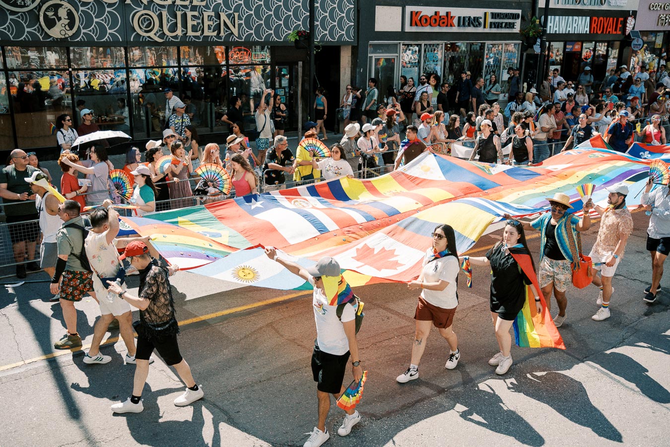 Participants in a parade carry a large flag featuring various national flags and rainbow colors, celebrating diversity and inclusion.