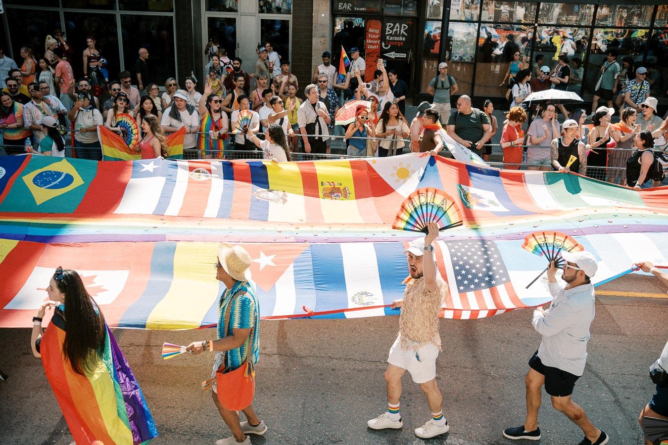 Participants in a parade carry a large flag displaying various national flags, celebrating diversity and unity, while the crowd cheers enthusiastically.