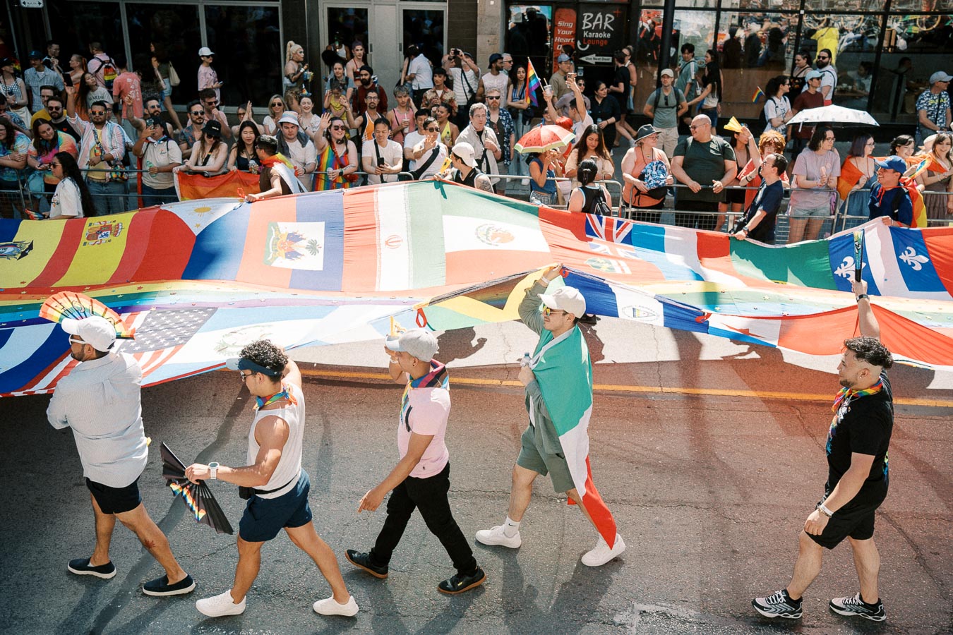 Participants in a parade carry a large flag featuring multiple national flags, while spectators watch from the sidelines.