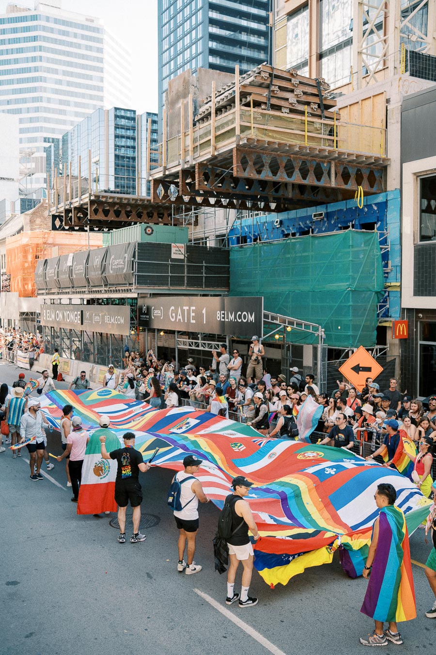 Participants in a parade hold a large flag featuring various national flags and rainbow colors, symbolizing diversity and inclusion, on a city street.