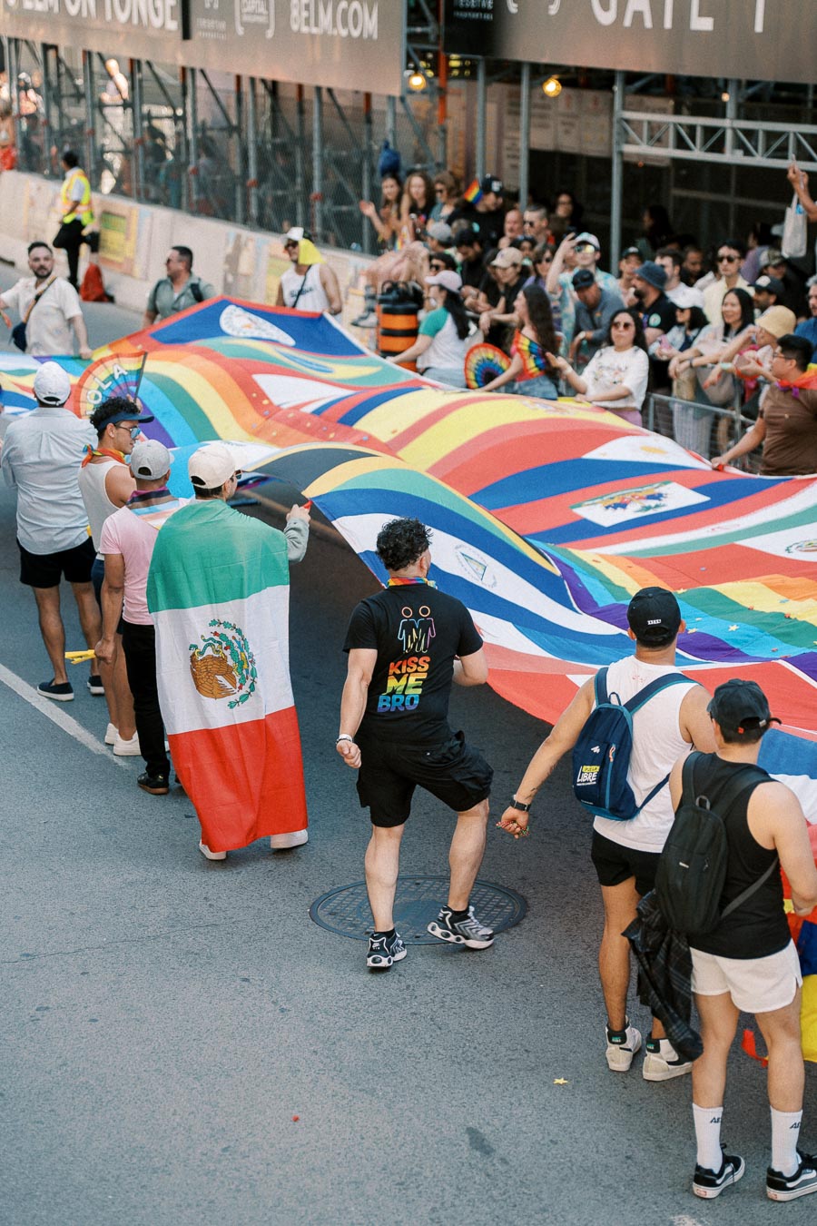 Participants in a pride parade hold a large rainbow flag adorned with various national flags, celebrating diversity and inclusion.