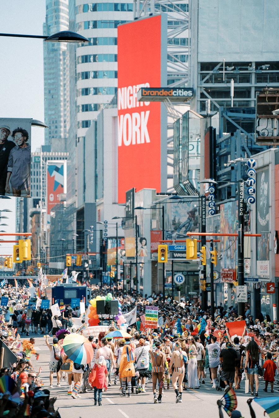 A lively parade takes place on a bustling city street, featuring participants in colorful costumes and rainbow flags, likely celebrating a pride event.