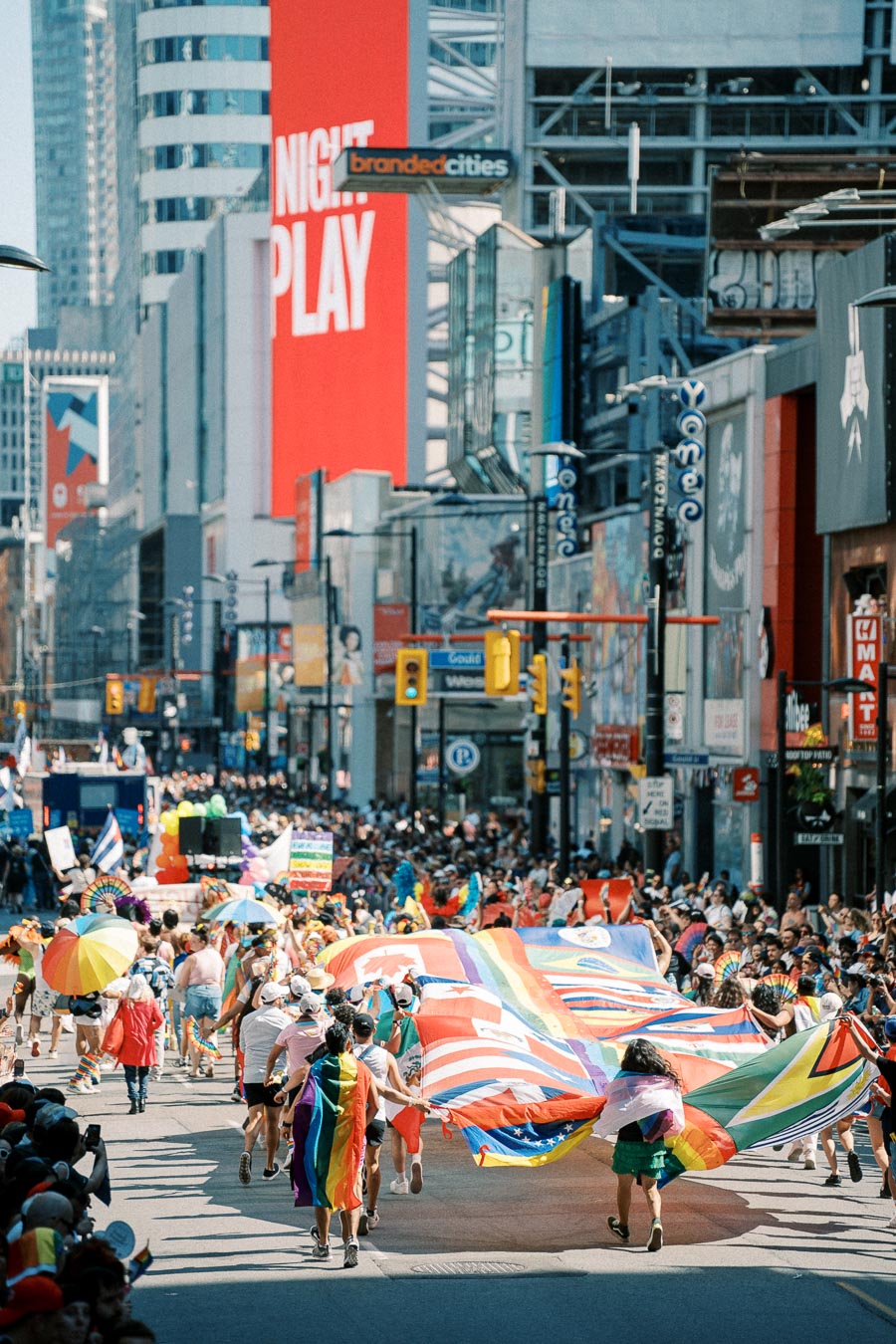 A lively parade featuring a large rainbow flag and various national flags, taking place in a bustling city street lined with tall buildings and colorful billboards.