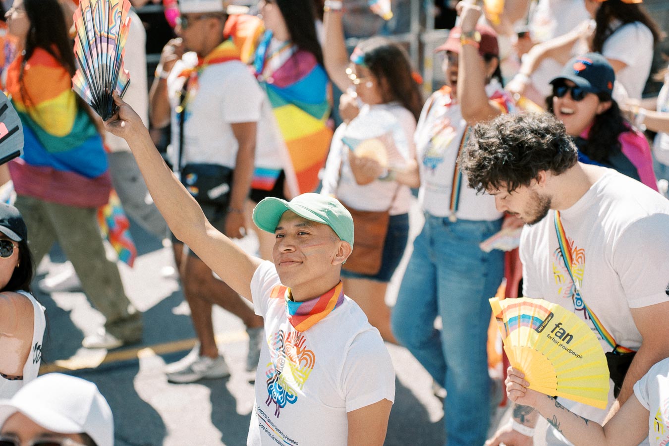 Participants at a pride parade celebrate with rainbow-themed clothing and accessories, including fans and scarves.