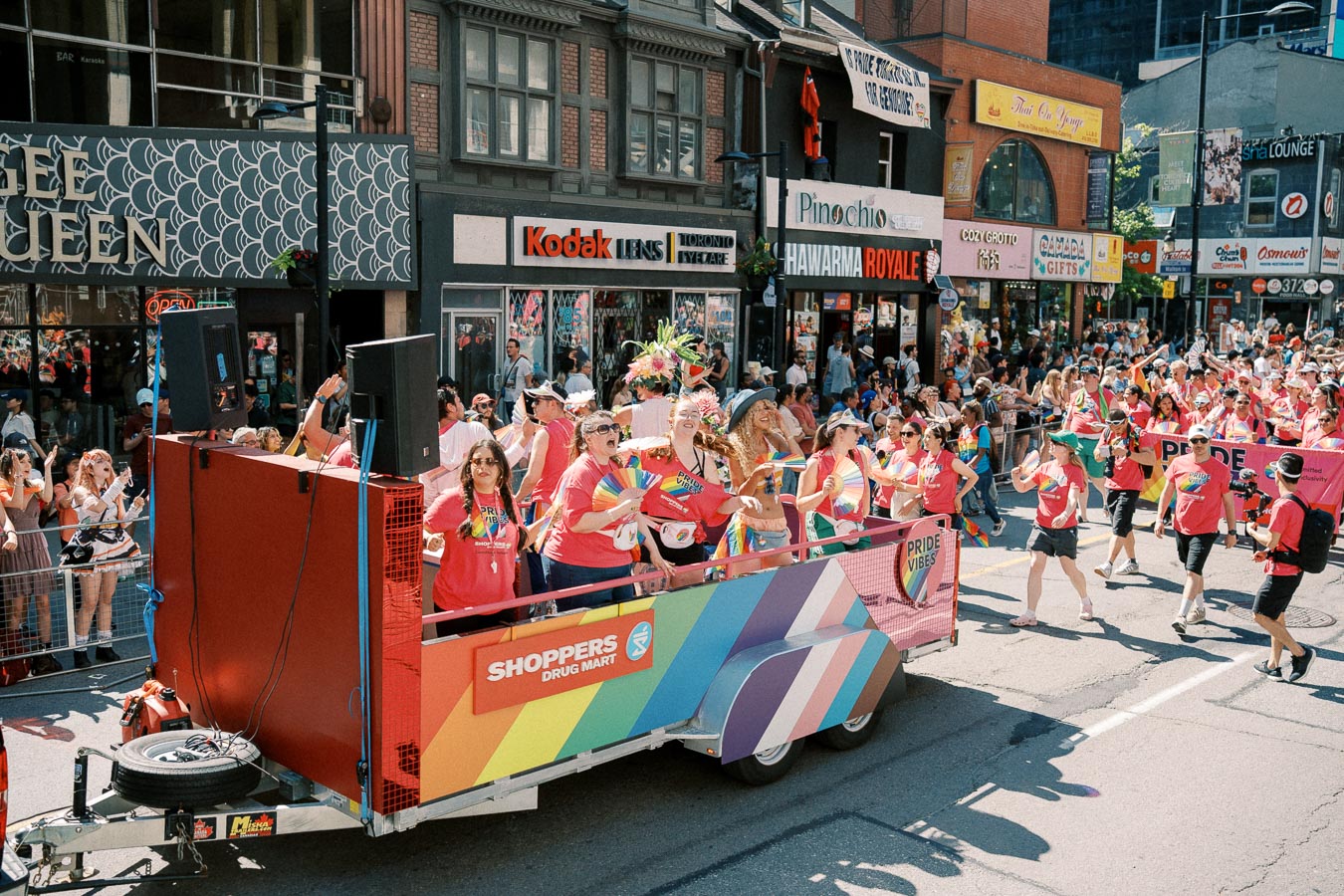 A lively scene from a Pride parade featuring a colorful float by Shoppers Drug Mart. Participants in pink shirts celebrate with rainbow flags, while spectators watch from the sidelines. The street is lined with various shops and restaurants.