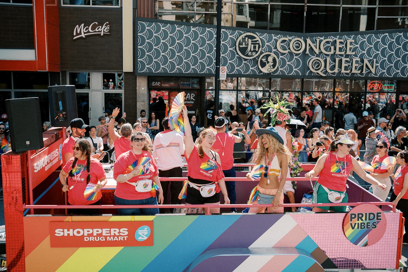 Participants celebrate on a colorful float during a Pride parade, showcasing vibrant rainbow decorations and signs for Shoppers Drug Mart and Pride Vibes. The event takes place in front of a Congee Queen restaurant, with a lively crowd in the background.