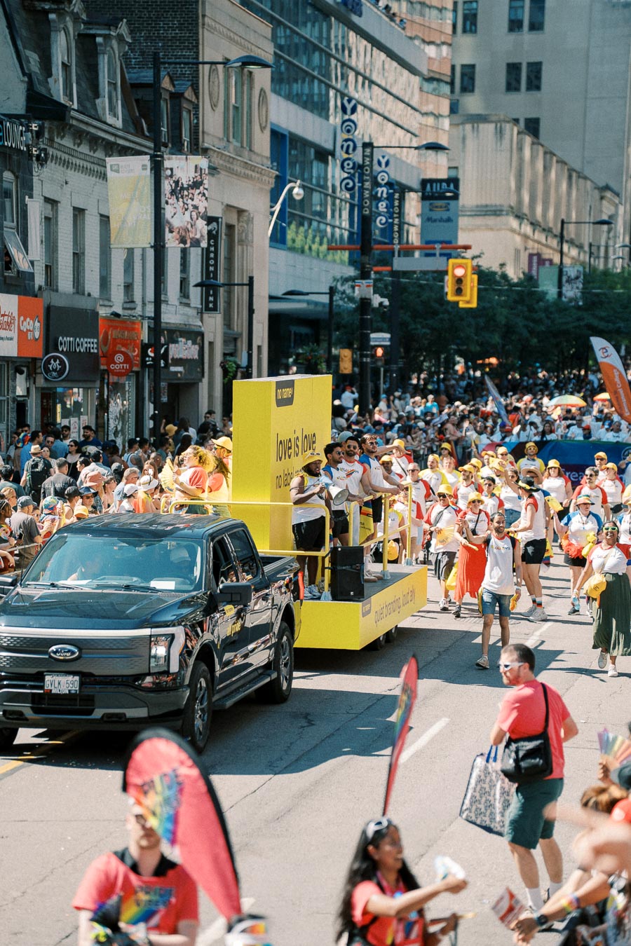 A lively parade featuring a yellow float with the message 'love is love' and participants in colorful attire, taking place on a bustling city street.