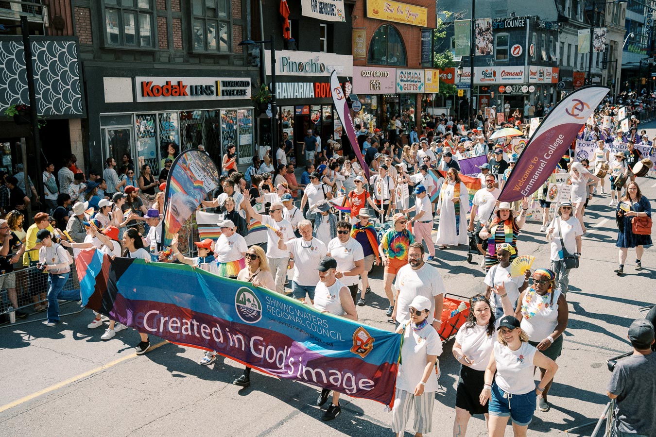 Participants in a pride parade march down a city street, holding colorful banners and flags, while spectators line the sidewalks.